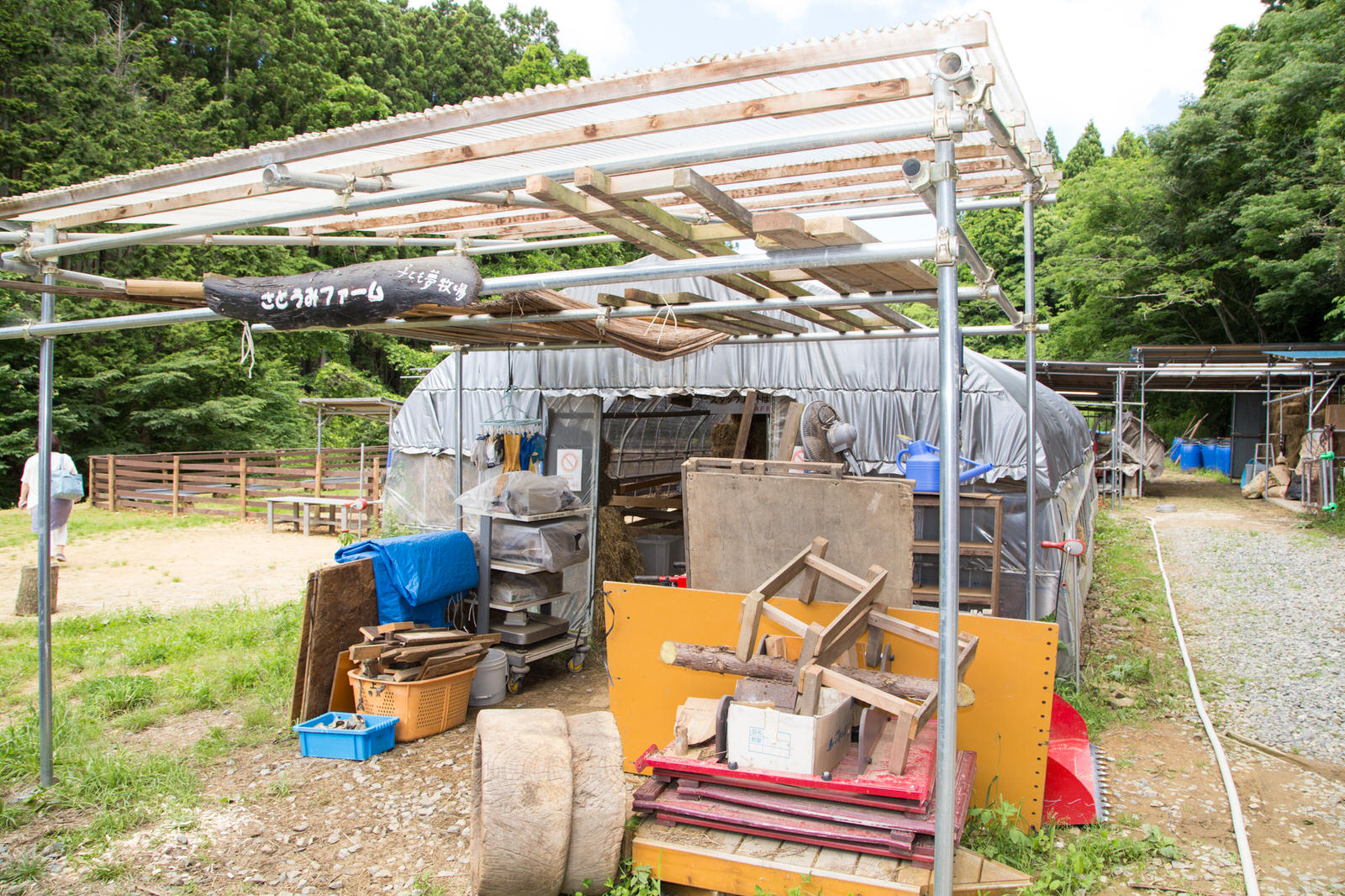 ビニールテントの手前に設置された鉄パイプの骨組みの屋根付きの作業場には、木材や棚などが雑然と置かれており、「さとうみファーム」という看板が掲げられている写真