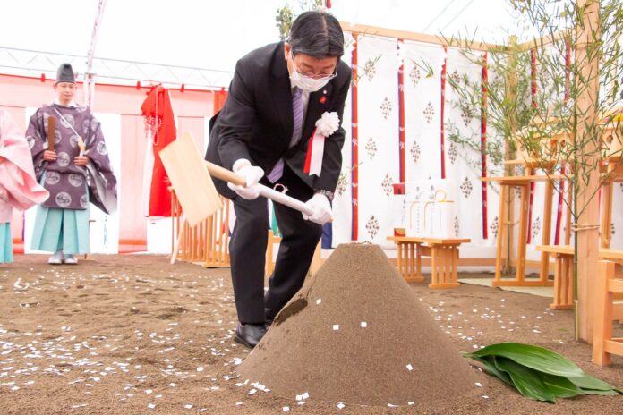 安全祈願祭の祭壇の前で、左胸に胸章リボンをつけ黒いスーツと白い手袋とマスクをした佐藤町長が、木製の鍬で盛り砂の山を掘り起している鍬入れの様子の写真