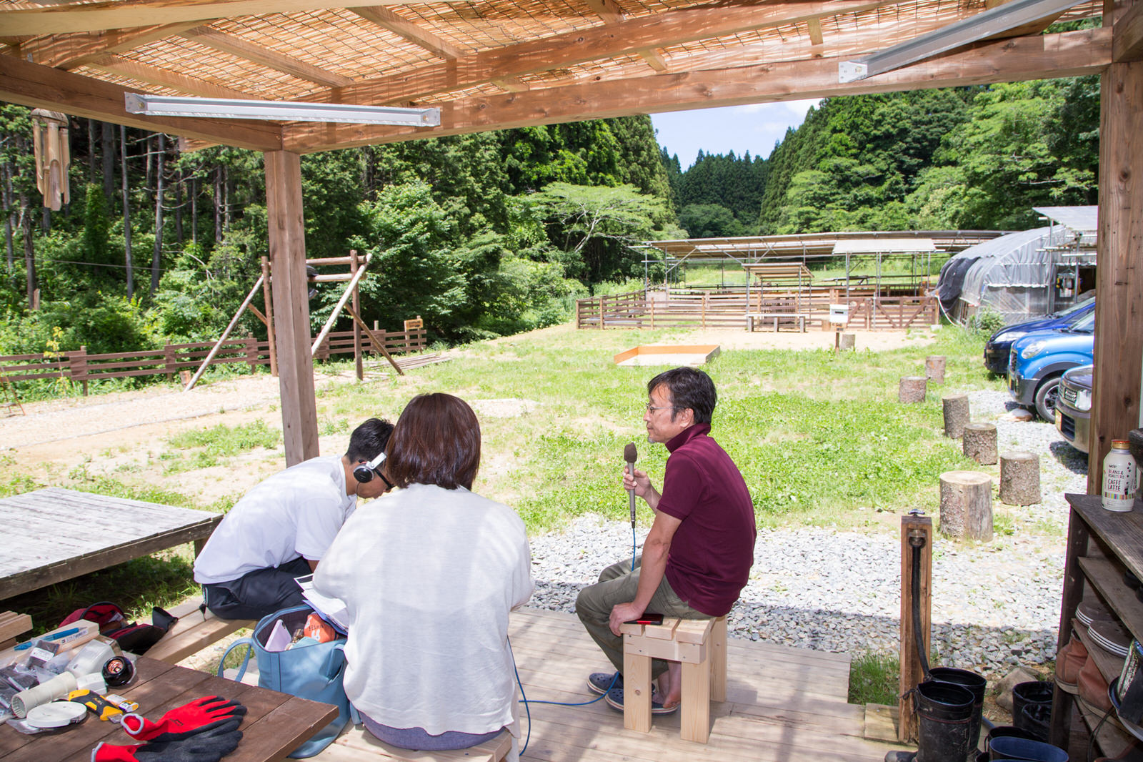 駐車スペース横にある屋根付きの作業場で、リポーターの女性と、ヘッドホンを着けた男性、マイクを持った金藤さんがそれぞれ木製のベンチに座っている写真