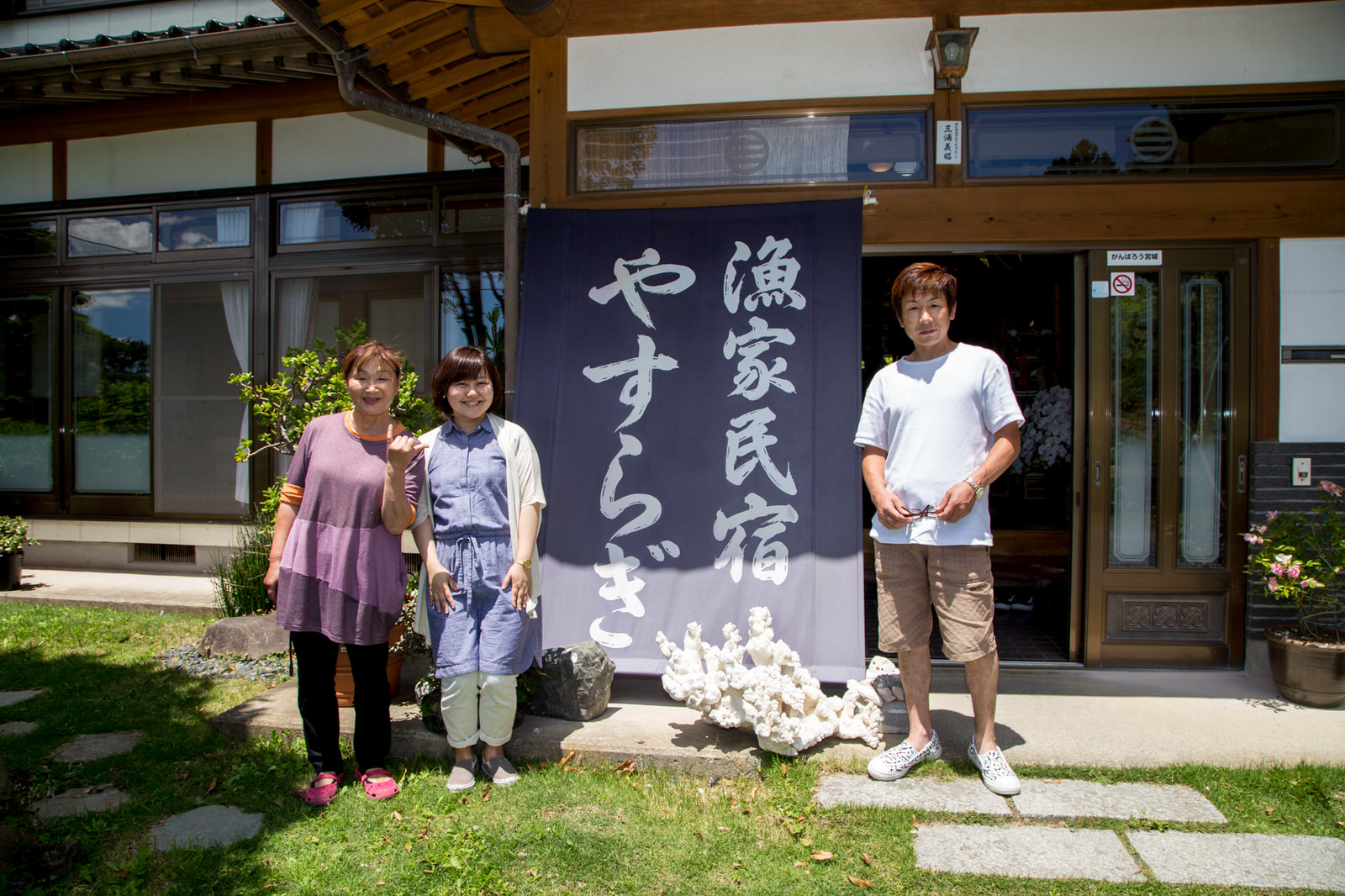 玄関先に「漁家民宿やすらぎ」と大きく書かれたのれんが掛かっていて、その前で笑顔の女性二人と男性一人が並んで立ち、晴天のもとであたたかく迎えている様子が写っている民宿の写真
