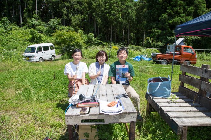 緑に囲まれた屋外のテーブルに書類やヘッドホンが置かれ、その奥にマイクを持った2人の女性と、パンフレットを持った男性が座っている写真