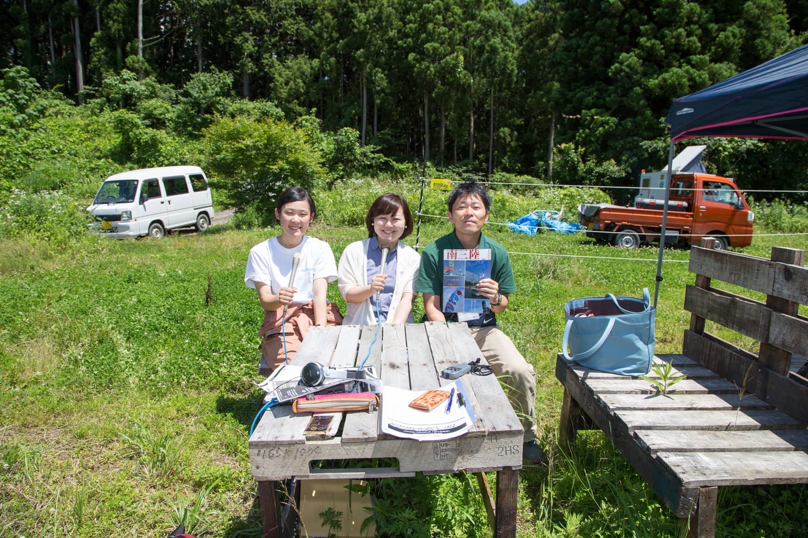 緑に囲まれた屋外のテーブルに書類やヘッドホンが置かれ、その奥にマイクを持った2人の女性と、パンフレットを持った男性が座っている写真