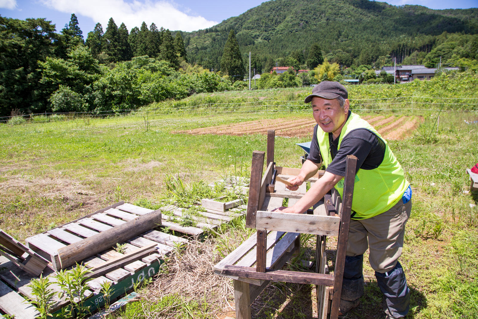 黄色いビブスを着た男性が、金づちを片手に、屋外で木材を使った作業をしており、後ろには畑や山など緑豊かな風景が広がっている写真
