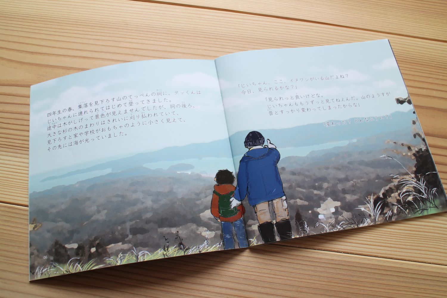 木製の机の上に、山の頂上から遠くの景色を見つめる男の子とおじいちゃんが描かれている絵本の中身が開いて置かれている写真