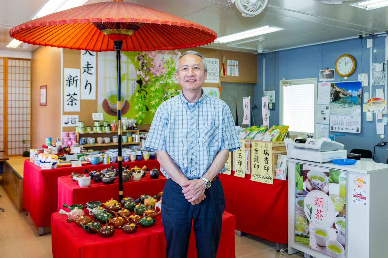 赤いテーブルクロスが敷かれた陳列棚に急須や湯飲みが並び、和傘が中央に建てられた和風の店内で男性店主が笑顔で立っている写真