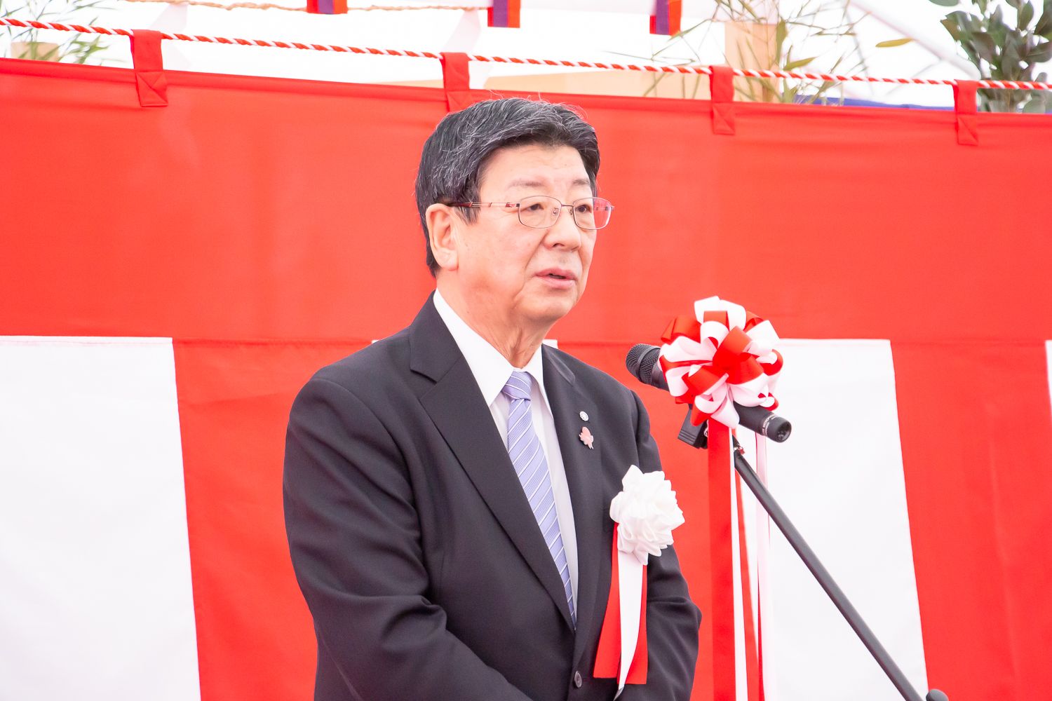 紅白幕が貼られた安全祈願祭の会場で、左胸に胸章リボンをつけ水色のネクタイを締めたスーツ姿の佐藤町長がマイクスタンドの前で挨拶を行っている式典の様子の写真