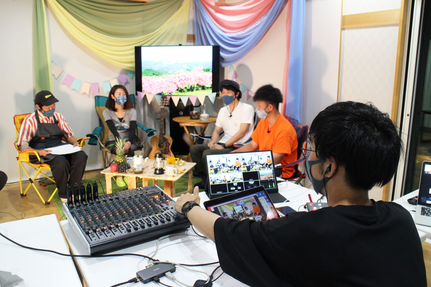 番組の収録現場で、手前に音響機器やスイッチャーを操作する男性が座っており、奥では5人がセットに座って談笑している様子が映っている写真