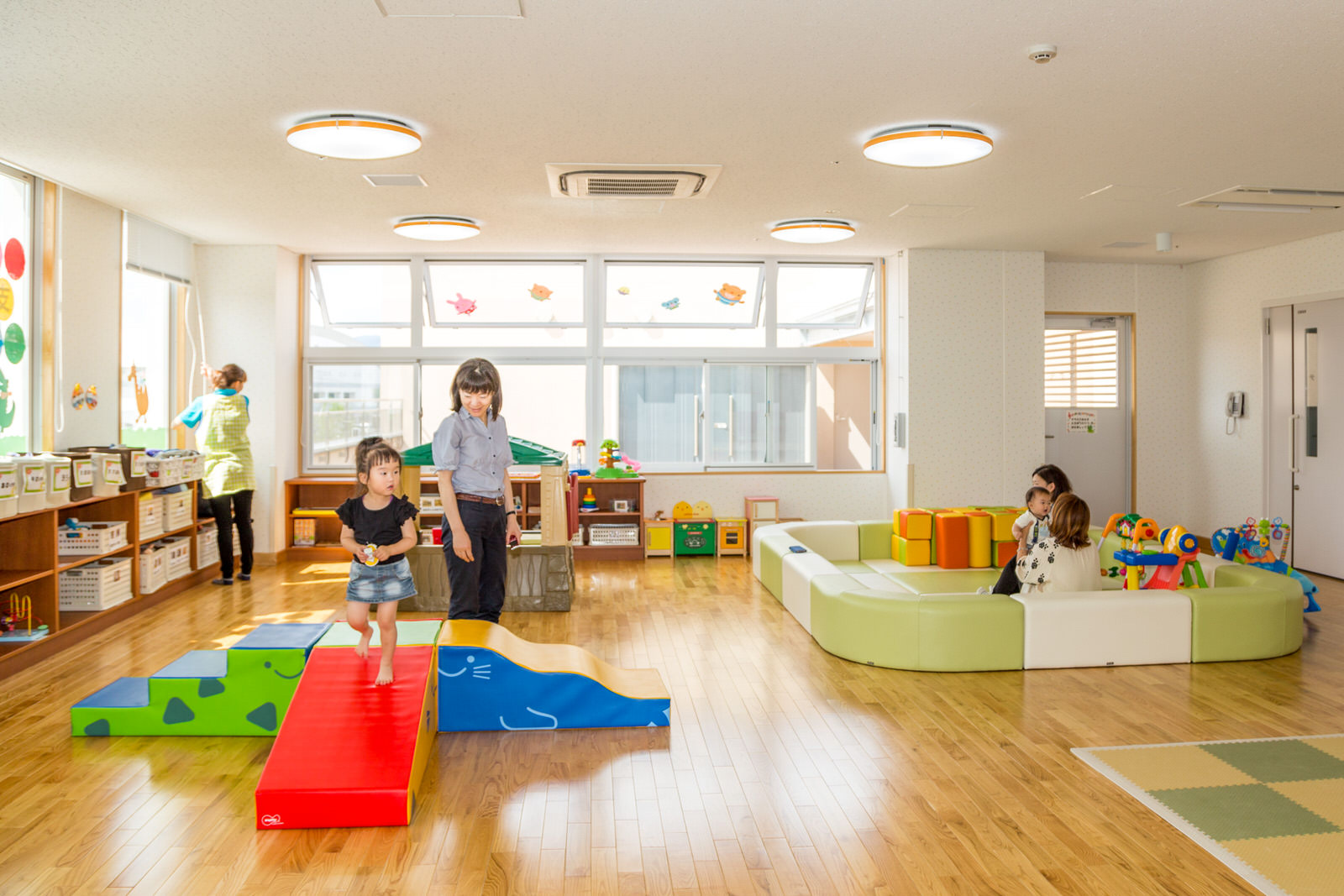明るい板張りの部屋の大きな窓の下におもちゃ箱が入った棚が置かれ、カラフルな遊具や小さな子供が遊ぶクッション敷きのスペースが設けられた地域子育て支援センターで親子が遊んでいる様子の写真