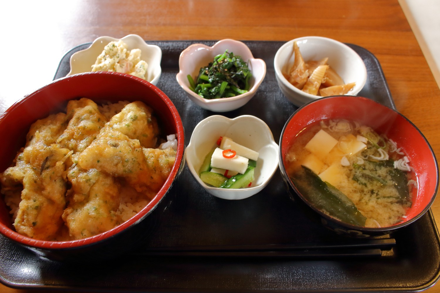 大きな赤い丼に盛り付けられたカキ天丼と、味噌汁、漬物、3つの小鉢が黒いお盆に乗せられているBランチの写真