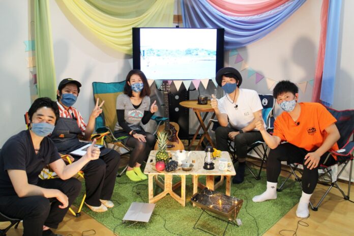 果物やランタンが置かれた中央のテーブルを囲んだ6人の男女がアウトドア風の装飾を背景にキャンプ用の椅子に座り、全員がマスクを着けて親しげにポーズをとっている写真