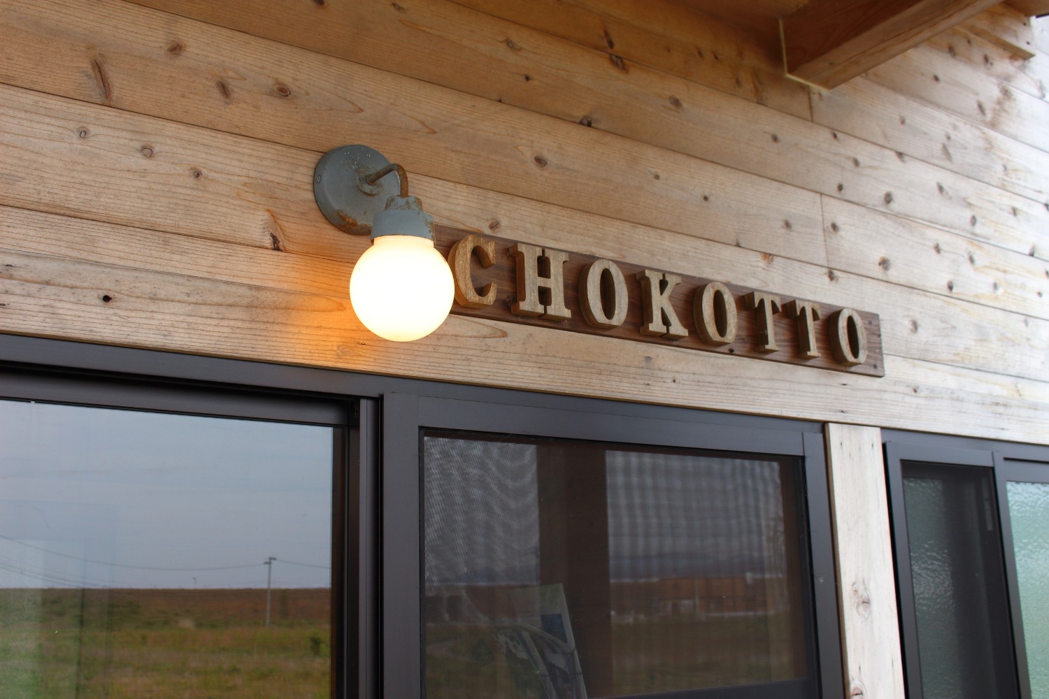 「CHOKOTTO」と書かれた木製の文字看板が玄関の上に取り付けられており、看板の左横に取り付けた照明の電球が温かく灯っている写真