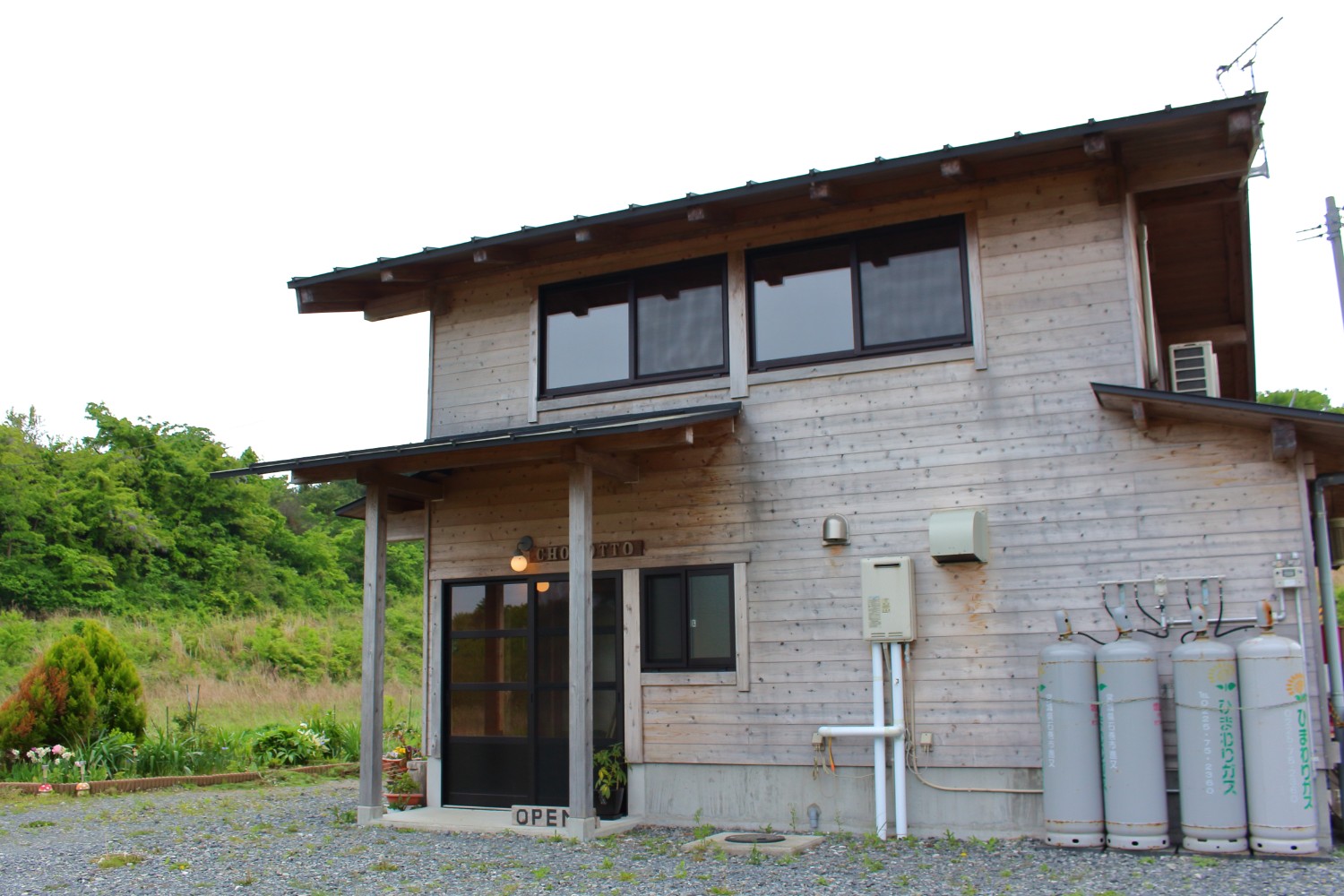 背景には花壇や草木が見える砂利の敷地に建つ、木造の二階建ての建物で入り口のドアの前にライトがついているカフェレストラン「ちょこっと」の外観写真