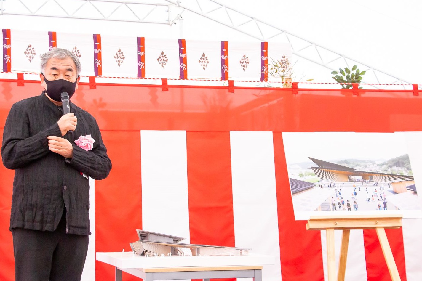紅白幕が貼られた安全祈願祭の会場の右側に近代的な建物の完成予想図が描かれたパネルがイーゼルに立てかけられ、中央のテーブルに建築模型が置かれ、左側にマイクを持つ隈研吾さんが立ち説明している写真