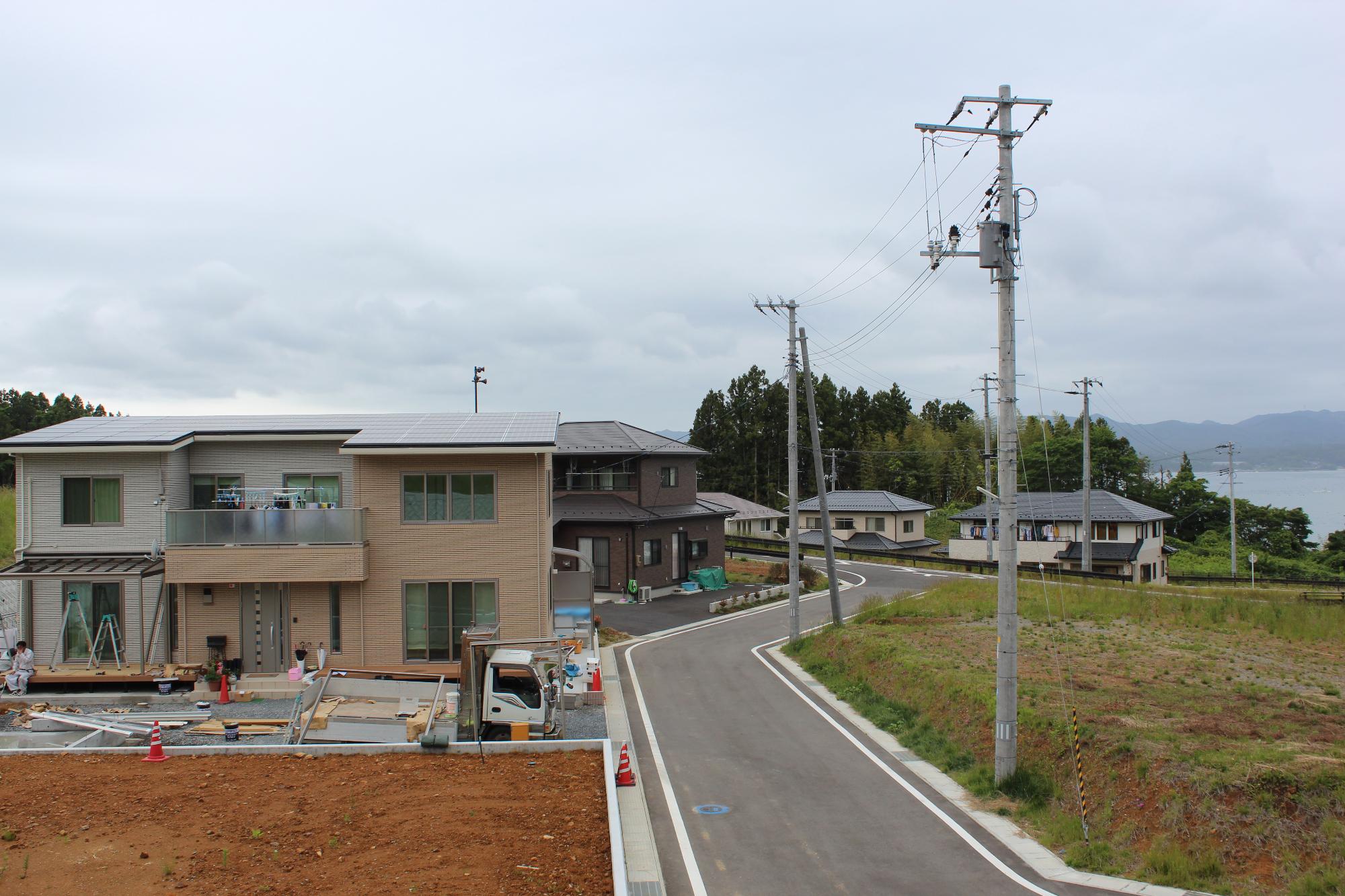 舗装された緩やかな坂道の脇に太陽光パネルを載せた新築の家が建ち、手前では外構工事が行われ、奥にはいくつもの住宅が並び、その向こうには海と山が広がる風景が見える写真