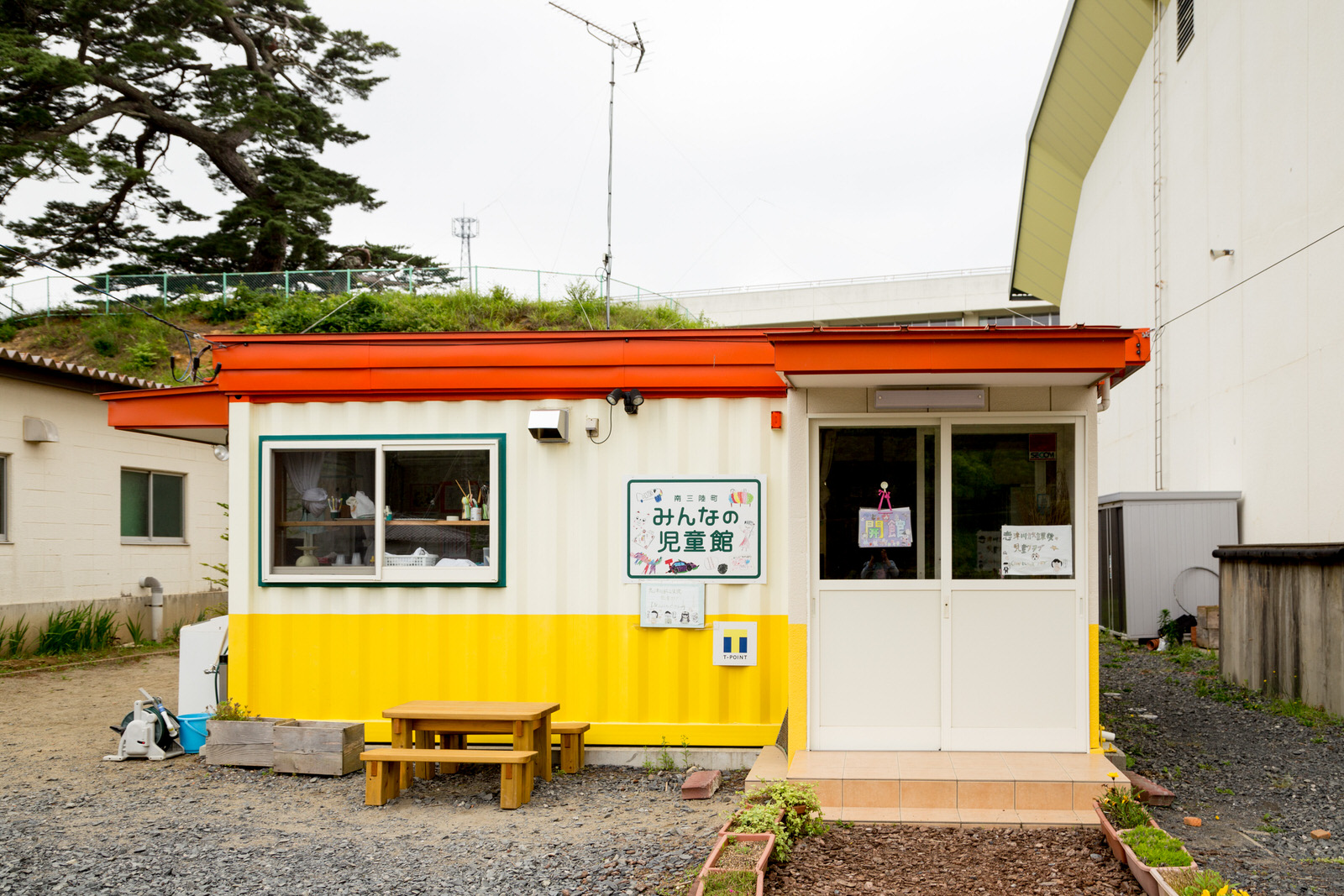 「みんなの児童館」の看板が設置された、オレンジ色の屋根と黄色と白の配色の壁のコンテナハウスに前に木製のベンチがある写真