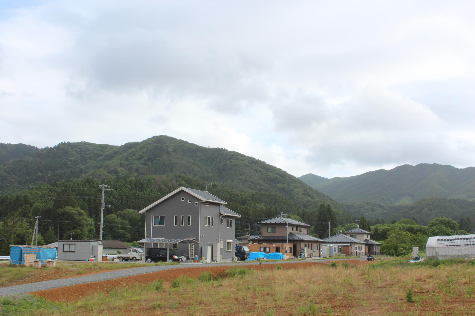 手前に赤土の畑と草地が広がり、その奥に新しい住宅が数軒建ち、背景には緑豊かな山が連なっている写真