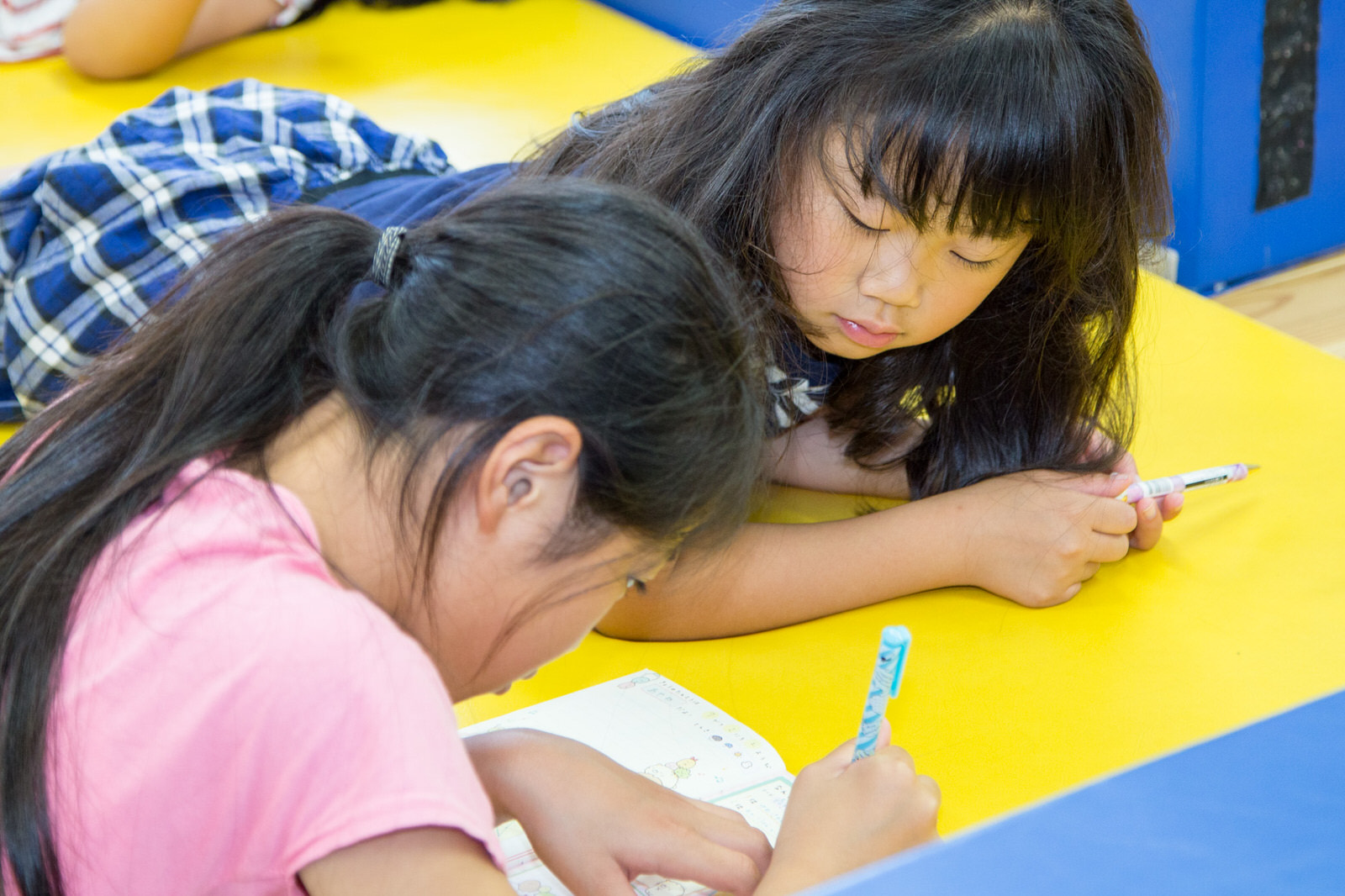 2人の女の子が黄色いマットの上に寝ころび、1人がノートに書きこんでいる様子をもう1人が眺めている様子の写真