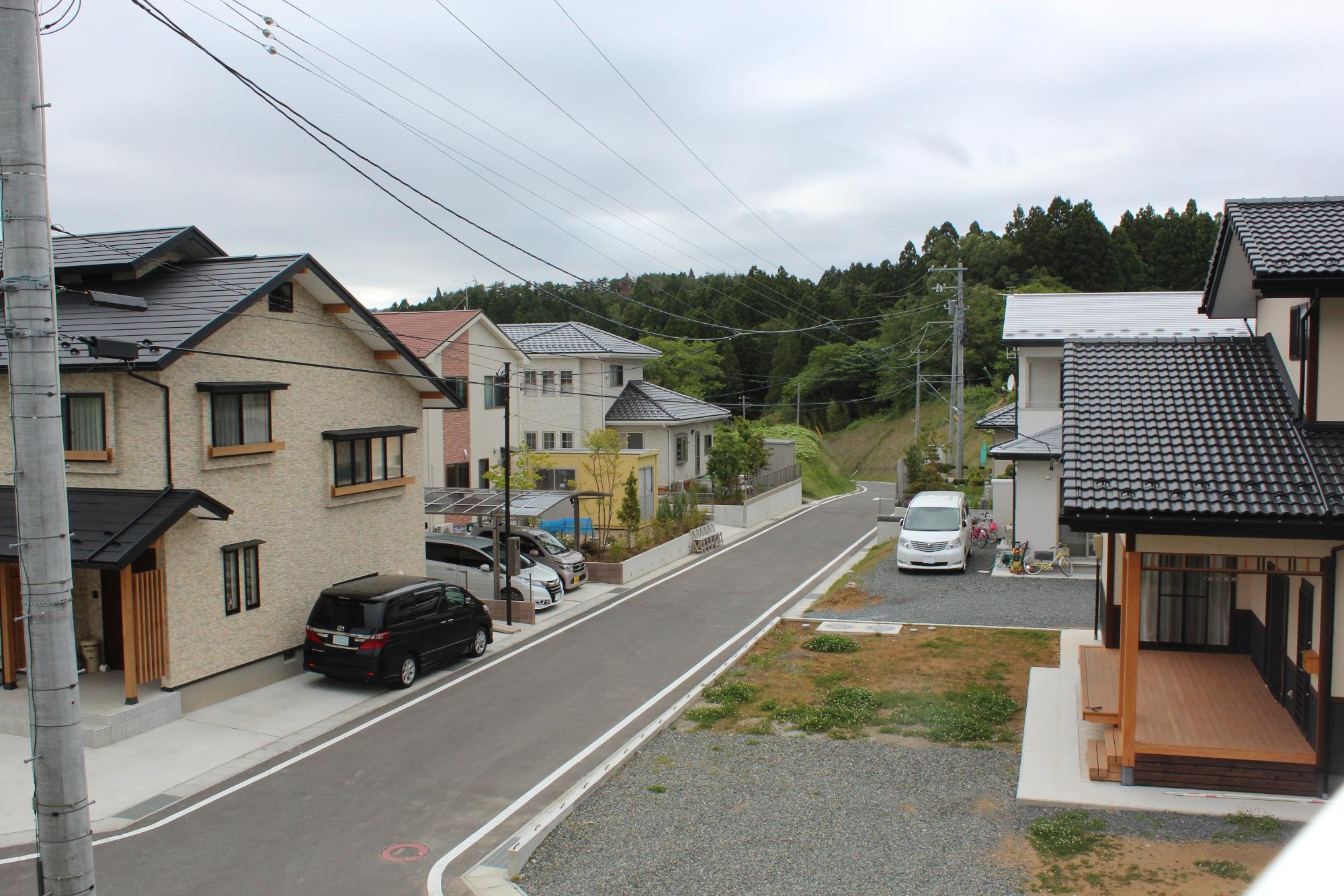 住宅街の一角で、舗装された道路の両側に新しい一軒家が並び、車が駐車され、背景には木々が広がる静かな住宅地の写真