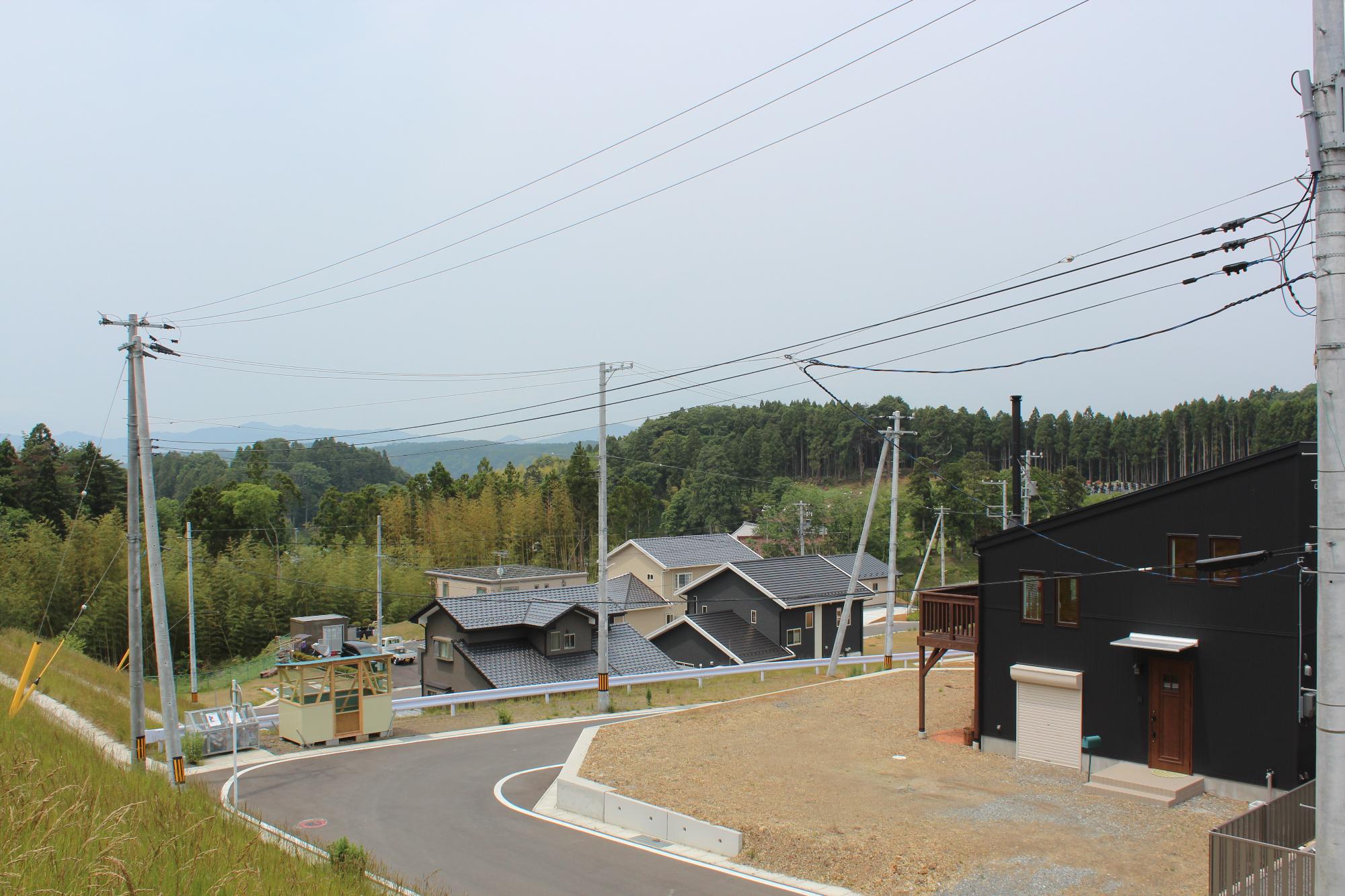 黒い外壁の新築住宅とその周囲にある数棟の住宅が、緑に囲まれた山間の住宅地に建ち並び、電柱と電線が交差している風景の写真