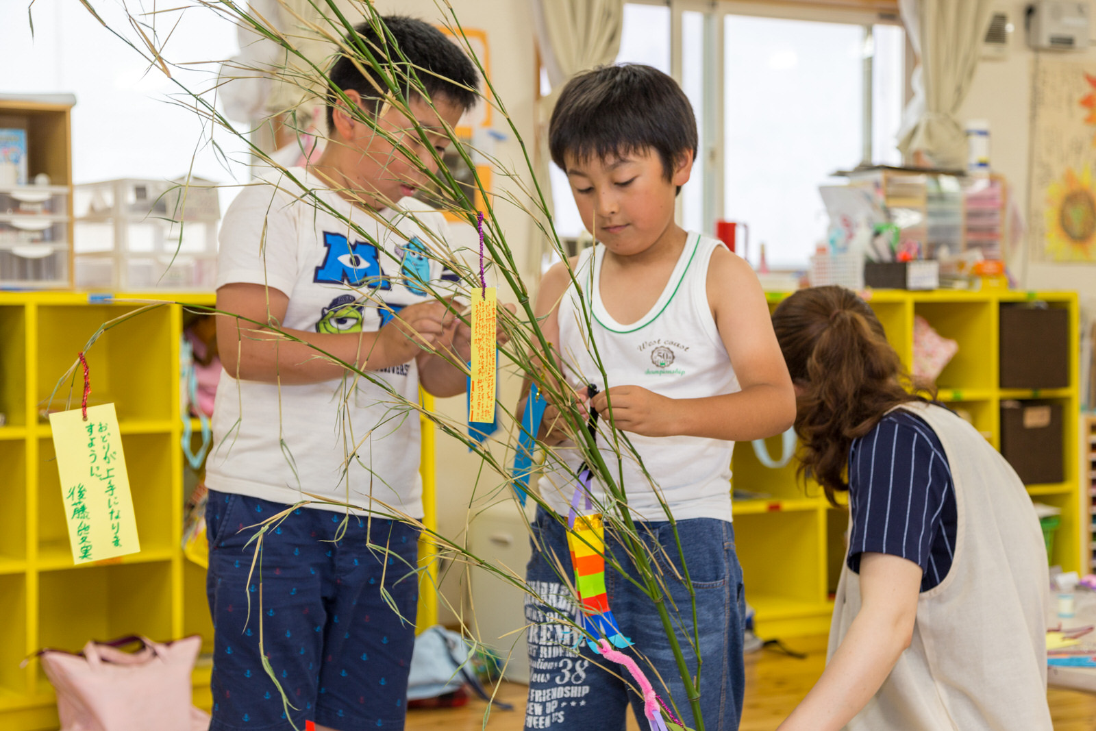 黄色い棚が設置された室内で、笹の枝に短冊を飾りつけている2人の男の子と、それを手伝う高橋さんの写真