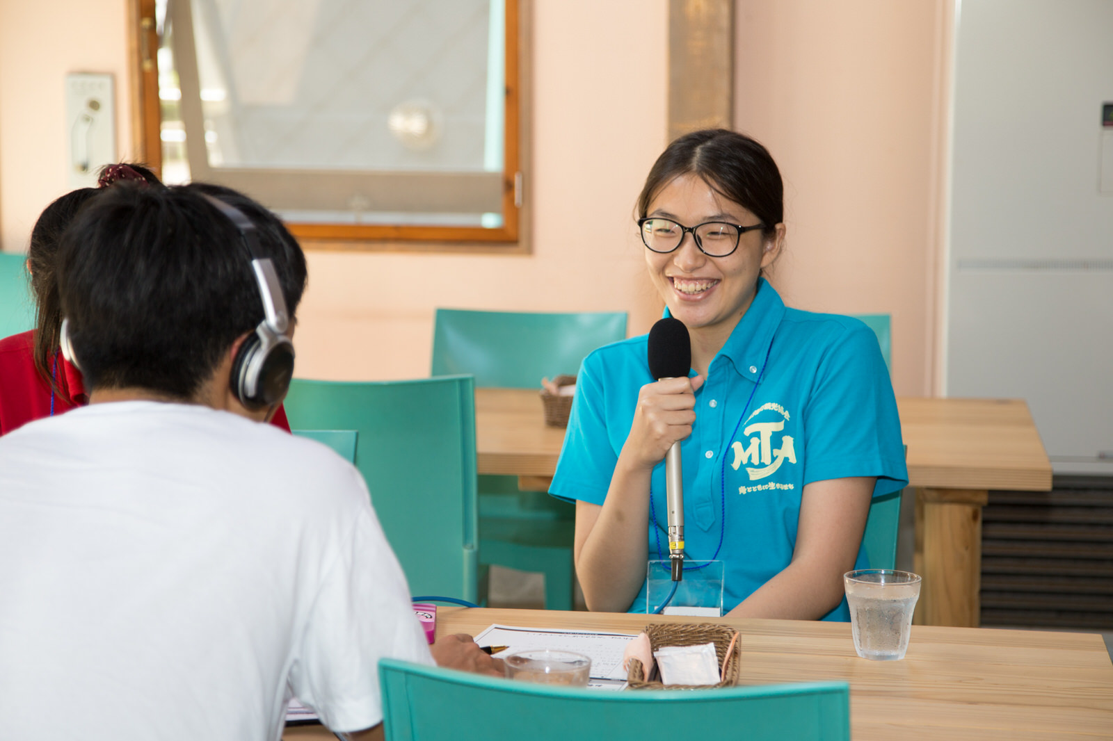 テーブルと水色の椅子が並んだ室内で、ヘッドホンをした男性とテーブルをはさんで座ったインターン生の女性が、マイクを持ち笑顔で取材を受けている様子の写真
