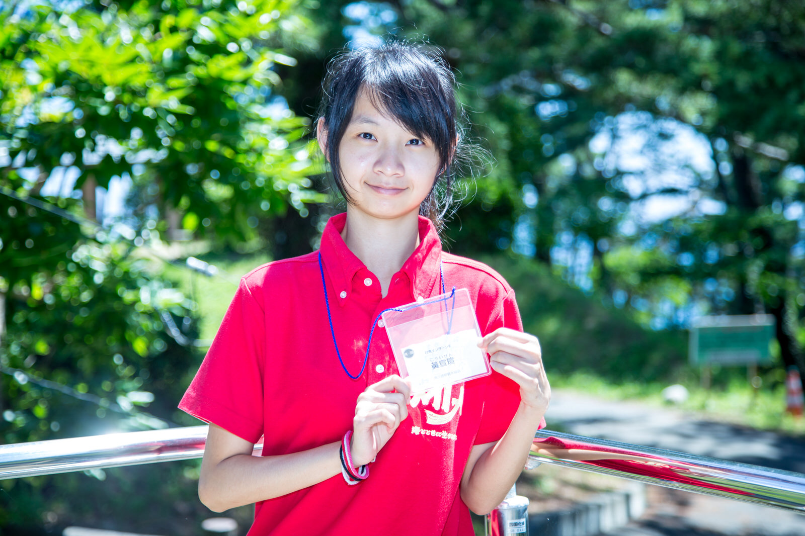 緑の木々を背景に、首からかけた名札を両手で胸の前に掲げ、笑顔でカメラを見ている女性の写真