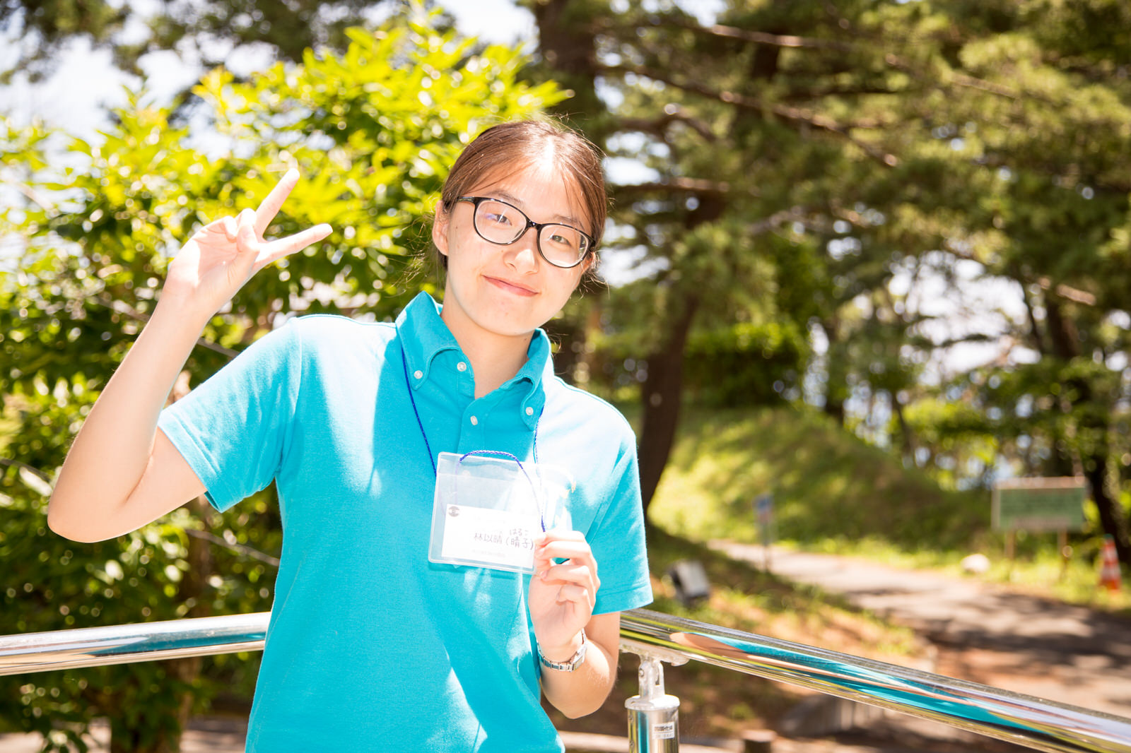 緑の木々を背景に、首からかけた名札を片手で胸の前に掲げ、笑顔でカメラにピースサインをしている女性の写真