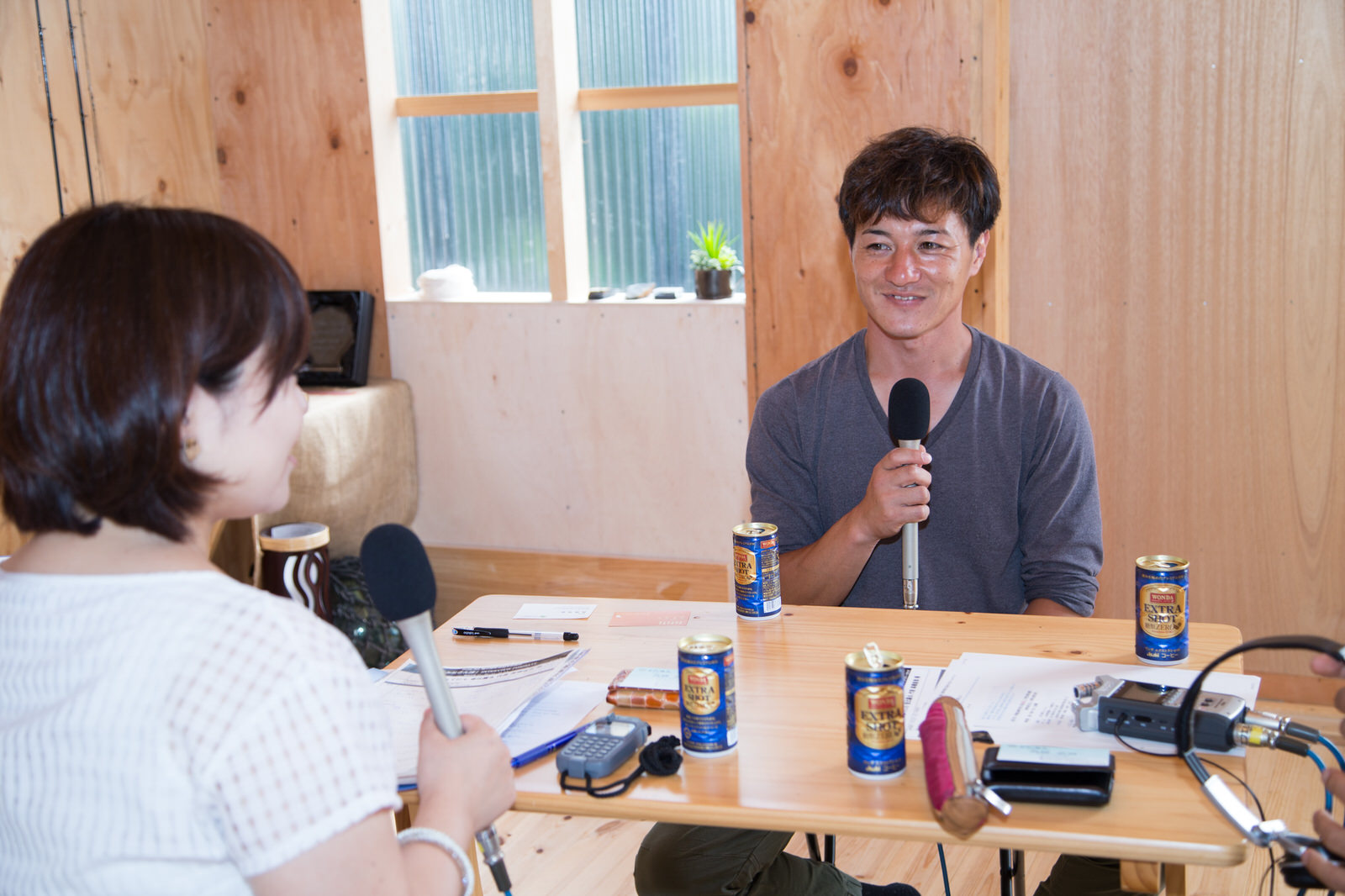 缶コーヒーや資料が置かれたテーブルをはさんで、リポーターの女性と高橋さんが向かい合って座り、マイクを持ってインタビューを行っている様子の写真