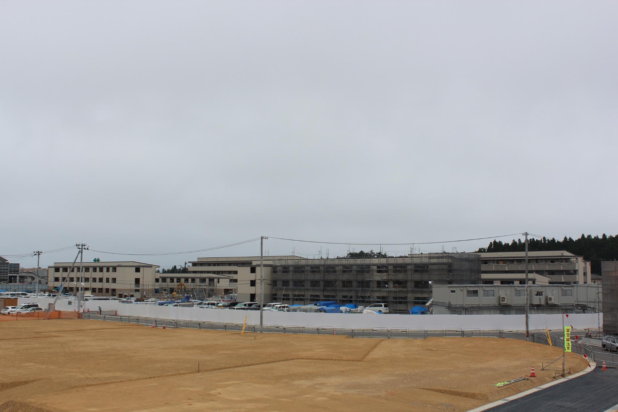 曇り空の下、仮囲いに囲まれた敷地内で建物の外観がほぼ完成し足場が一部残る中、複数の車両が並び整備が進む様子が見られる建設現場の写真