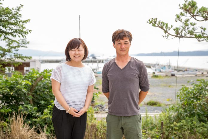 遠くに山影が見える海を背景に、木々に囲まれて並んで立っている、女性リポーターと高橋さんの写真