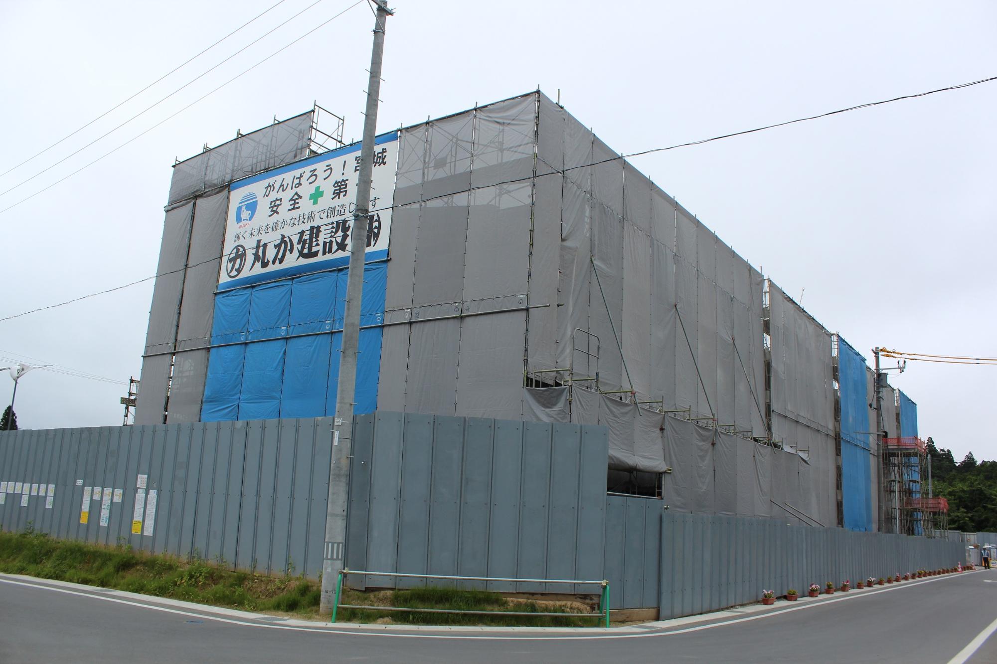 建設中の大規模な建物が灰色と青色の防音シートで覆われ、手前には安全標語入りの大きな看板が掲げられた工事現場の様子を示す写真