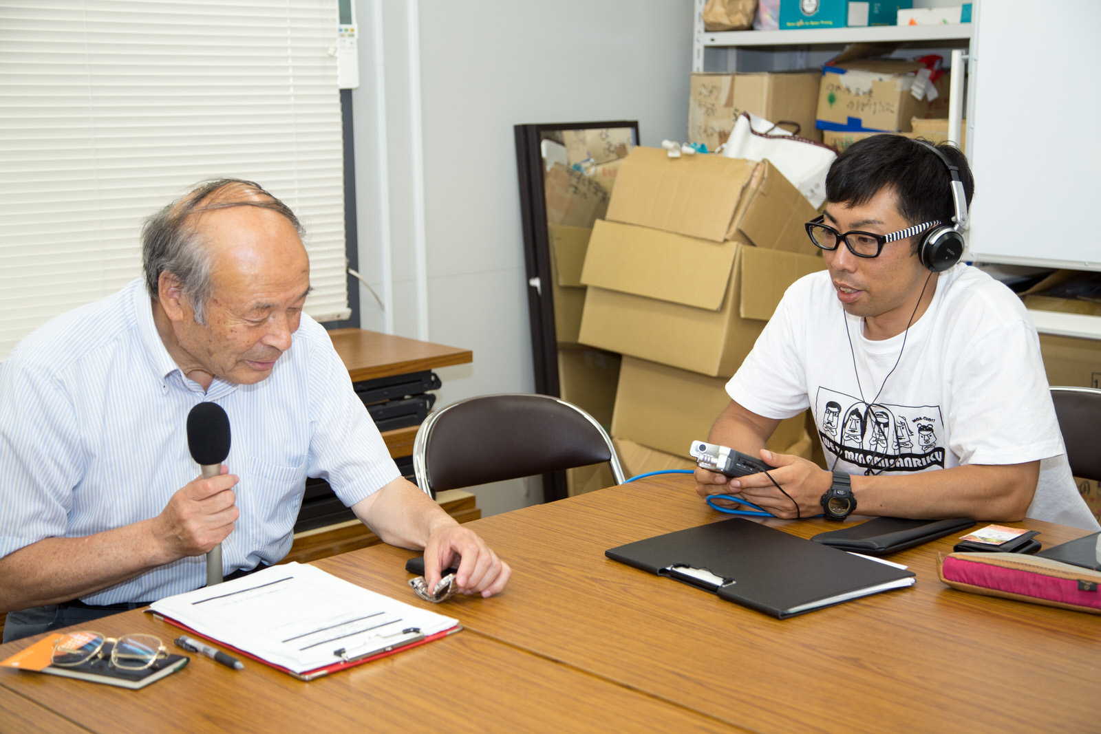 資料を置いたテーブルに座って、マイクを手に話をする千葉さんと、ヘッドホンを着けて機材を持った男性の写真