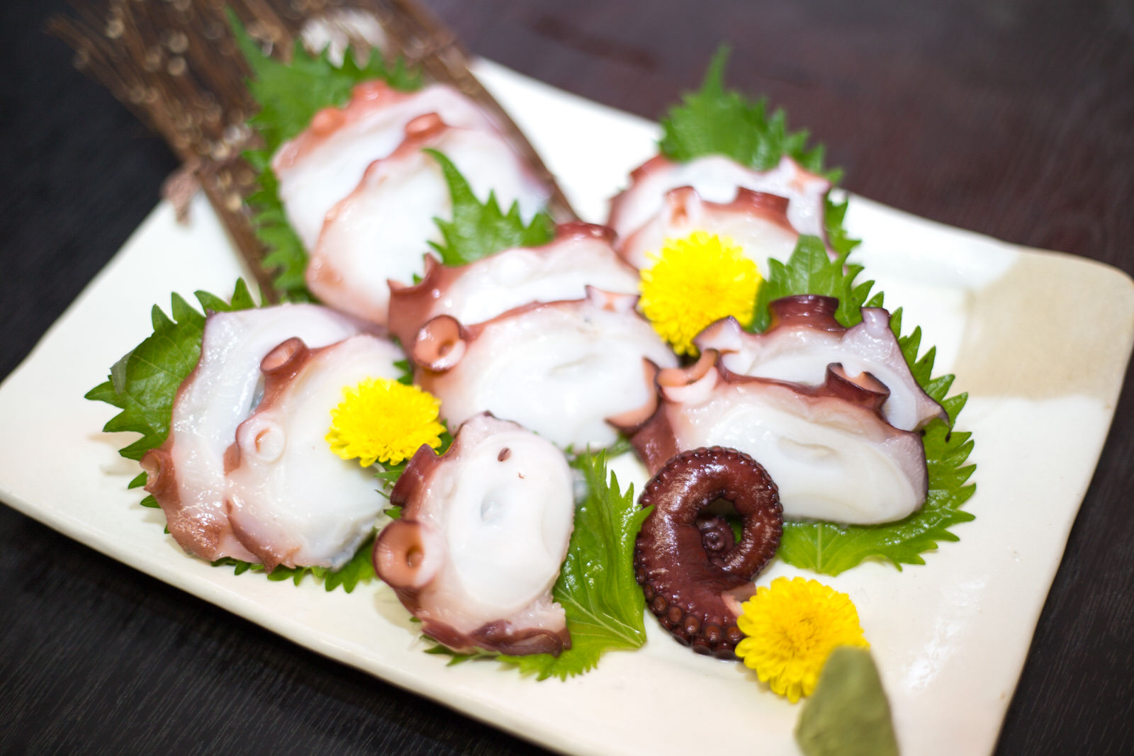 白い角皿に大きなタコの刺身が大葉や菊の花と一緒に美しく盛り付けられ、わさびが添えられている写真