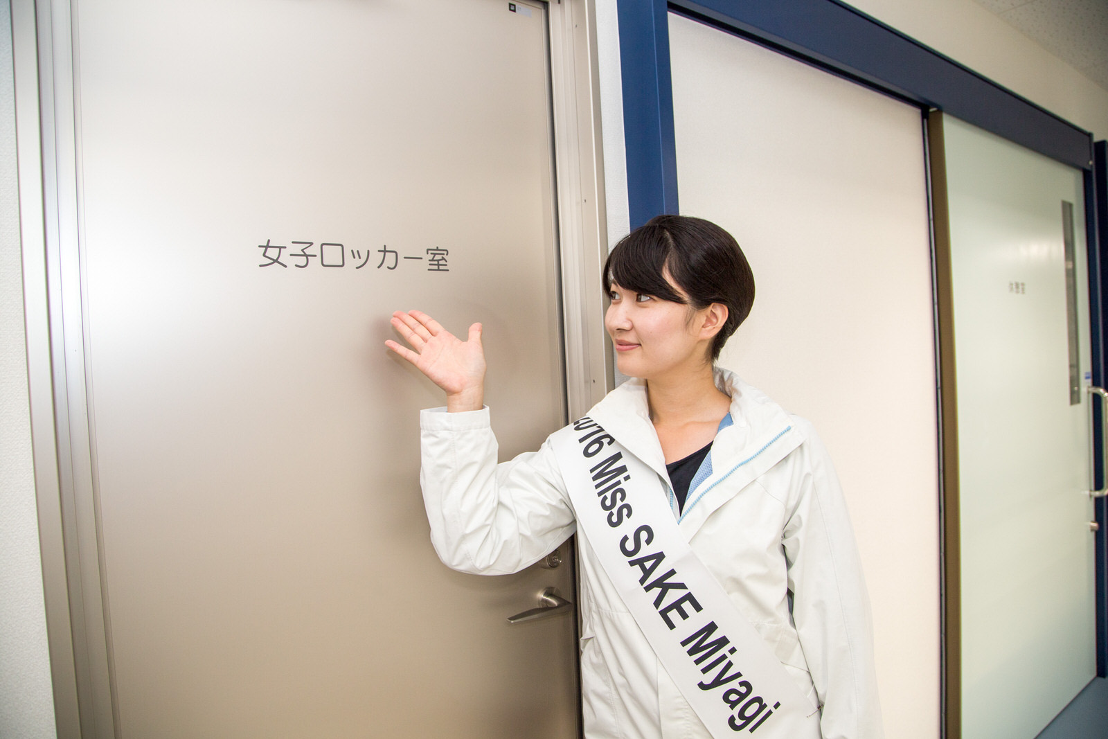 「女子ロッカー室」と書かれたベージュの扉の横に立った池田かほりさんが、ドアに書かれた文字を示している写真