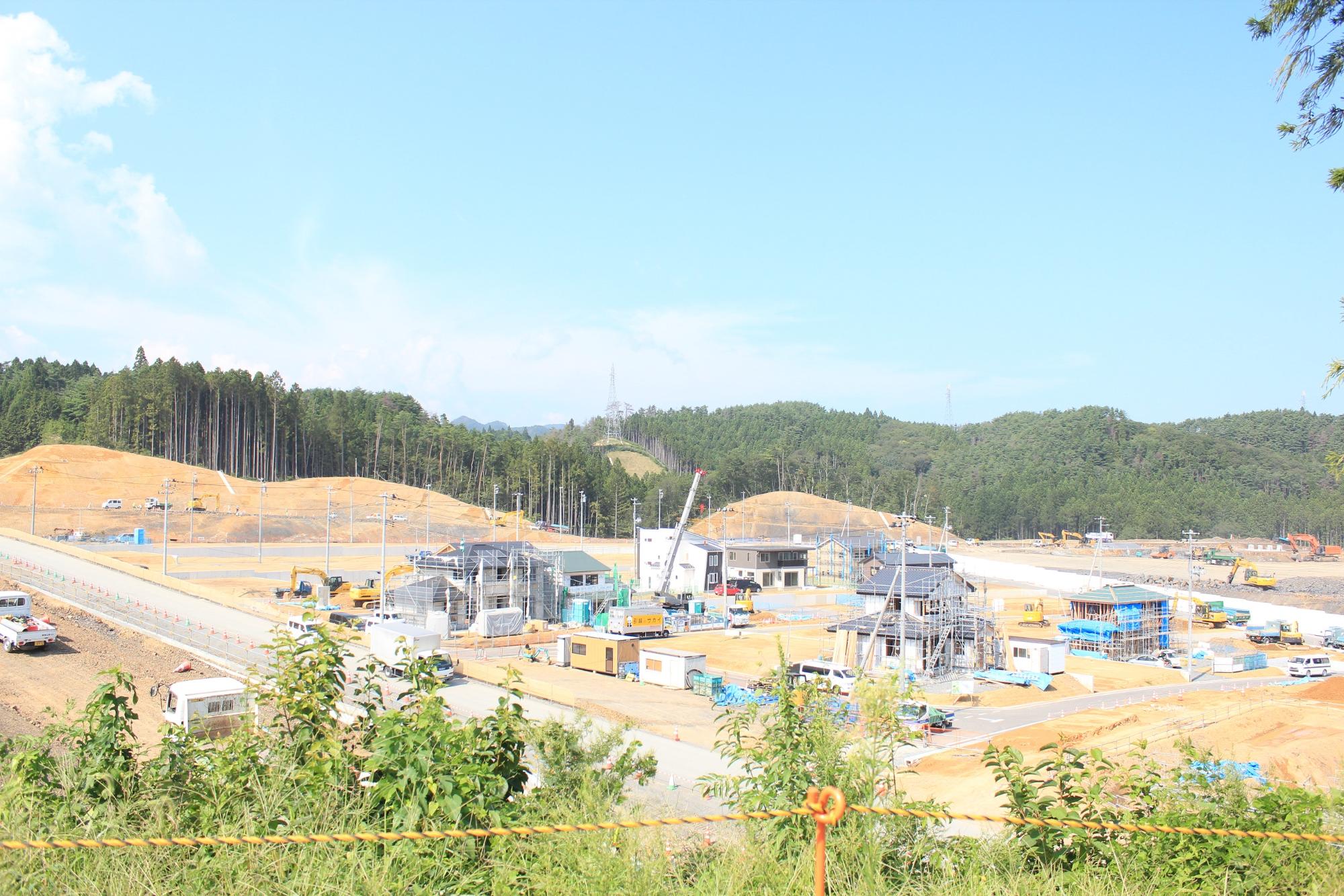 奥に緑の山林、手前に住宅区画があり、住宅区画では複数の家が建ち始め、右奥ではショベルカーなどの重機が工事を行っている様子が捉えられる写真