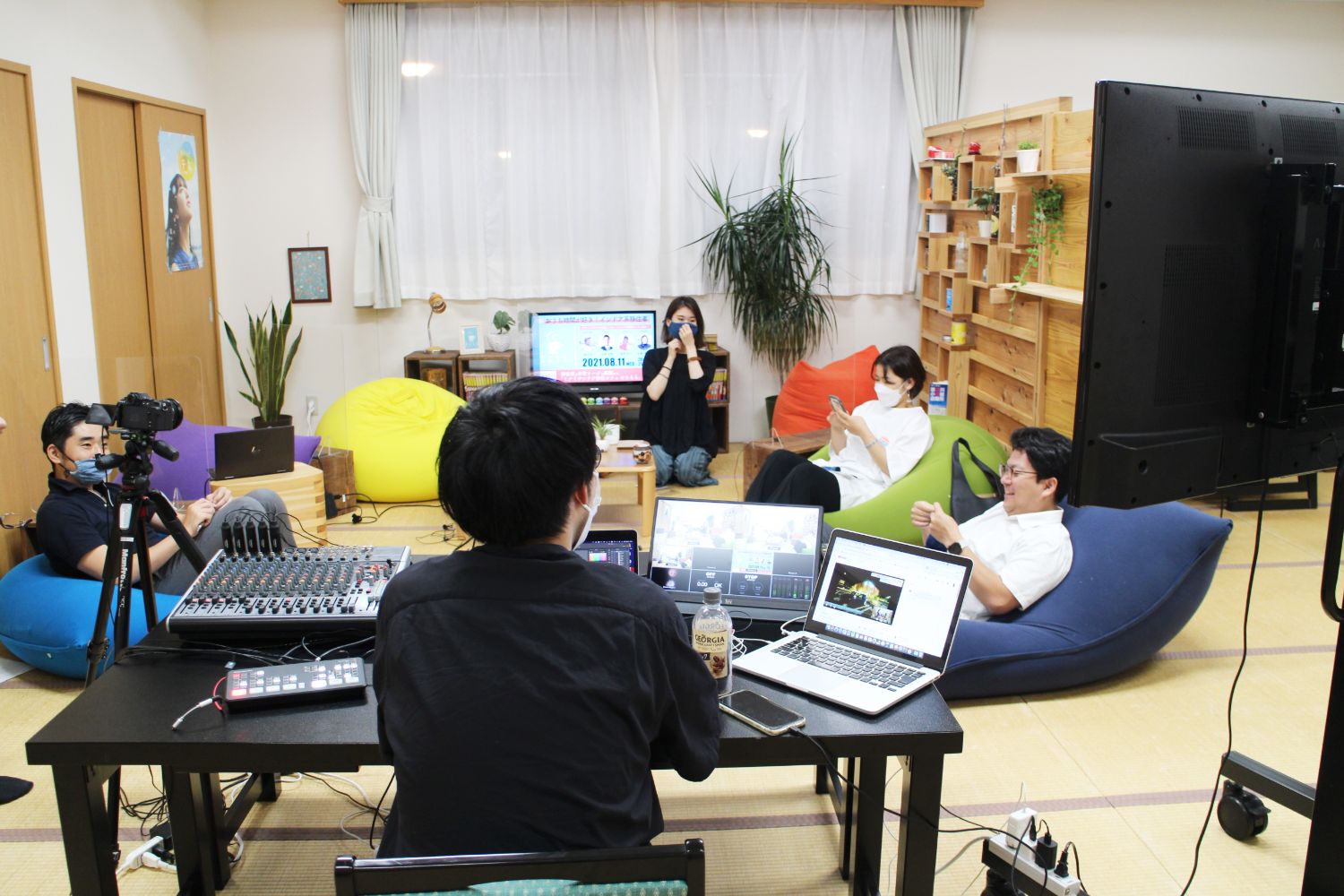 パソコンやカメラ、ミキサーなどが置かれた黒いテーブルに音響・映像機材を操作する男性が座り、畳の部屋でビーズクッションに座る4人の男女が写っている写真