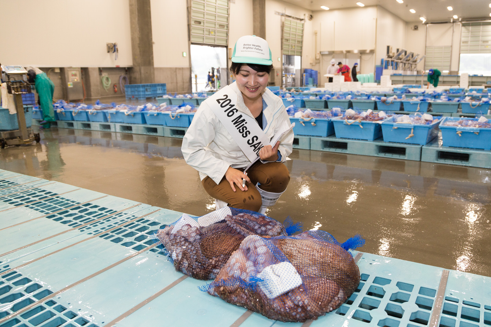 背後に魚介類が入った青いコンテナがたくさん並んだ市場内で、池田かほりさんがしゃがんで水色のパレットに2つ乗った青い網袋入りのタコを見ている写真