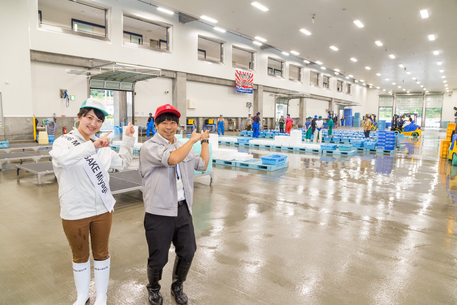 池田かほりさんと赤い帽子をかぶった西條さんが、立って並び、後ろに広がる広々とした明るい荷捌き場内を指差している写真