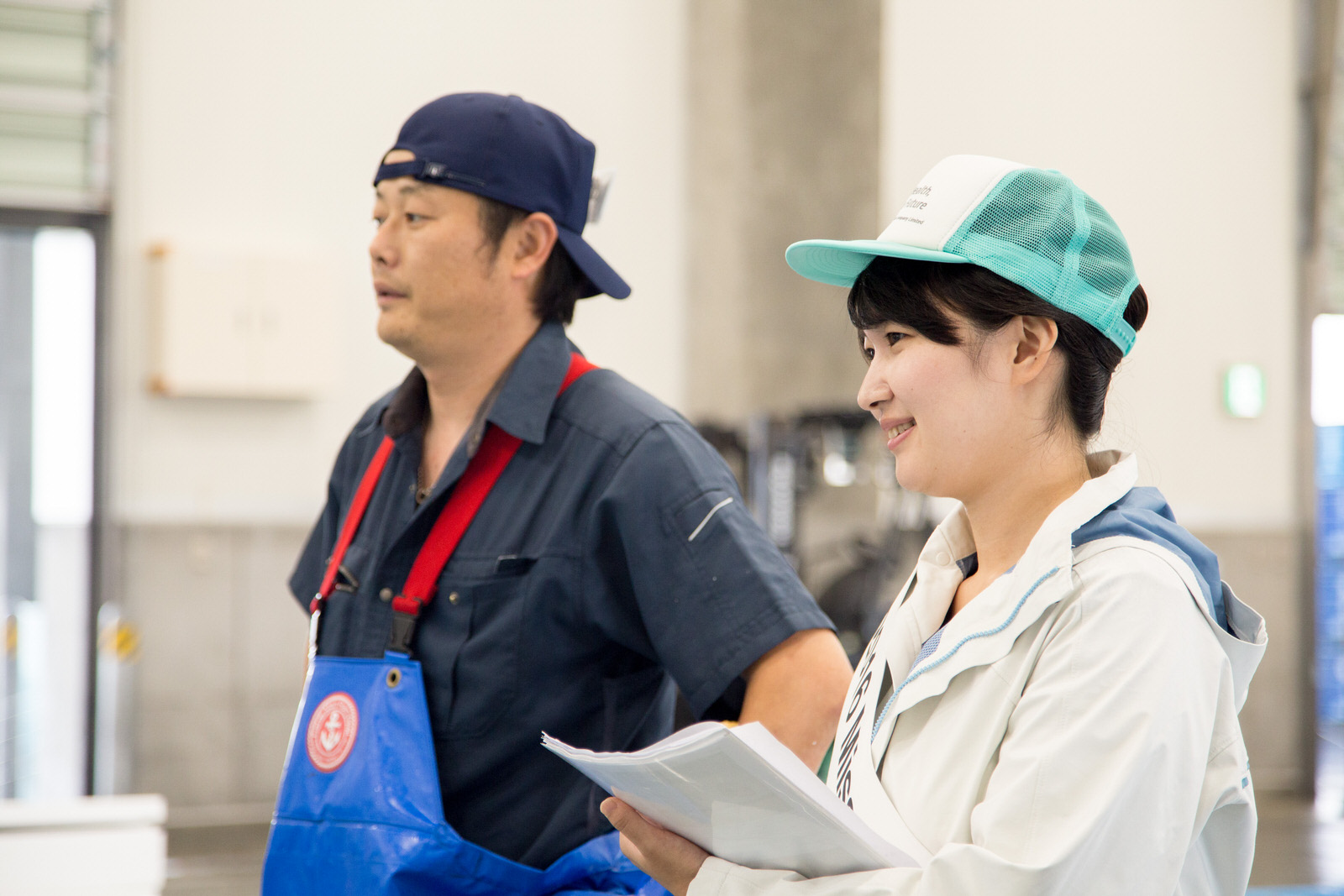 紺色の帽子をかぶった市場の関係者の男性と、水色の帽子をかぶり微笑んだ池田さんが並んで立っている様子を斜め右から写した写真