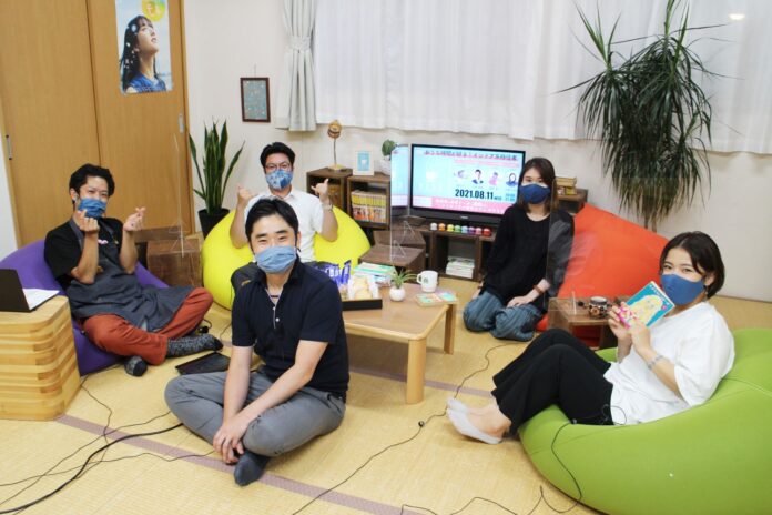 観葉植物、木の家具、テレビ、飲み物やお菓子が置かれたテーブルが置かれた畳敷きの部屋で、5人の男女がカラフルなビーズクッションに座り、青いマスクを着けてカメラを見ている写真