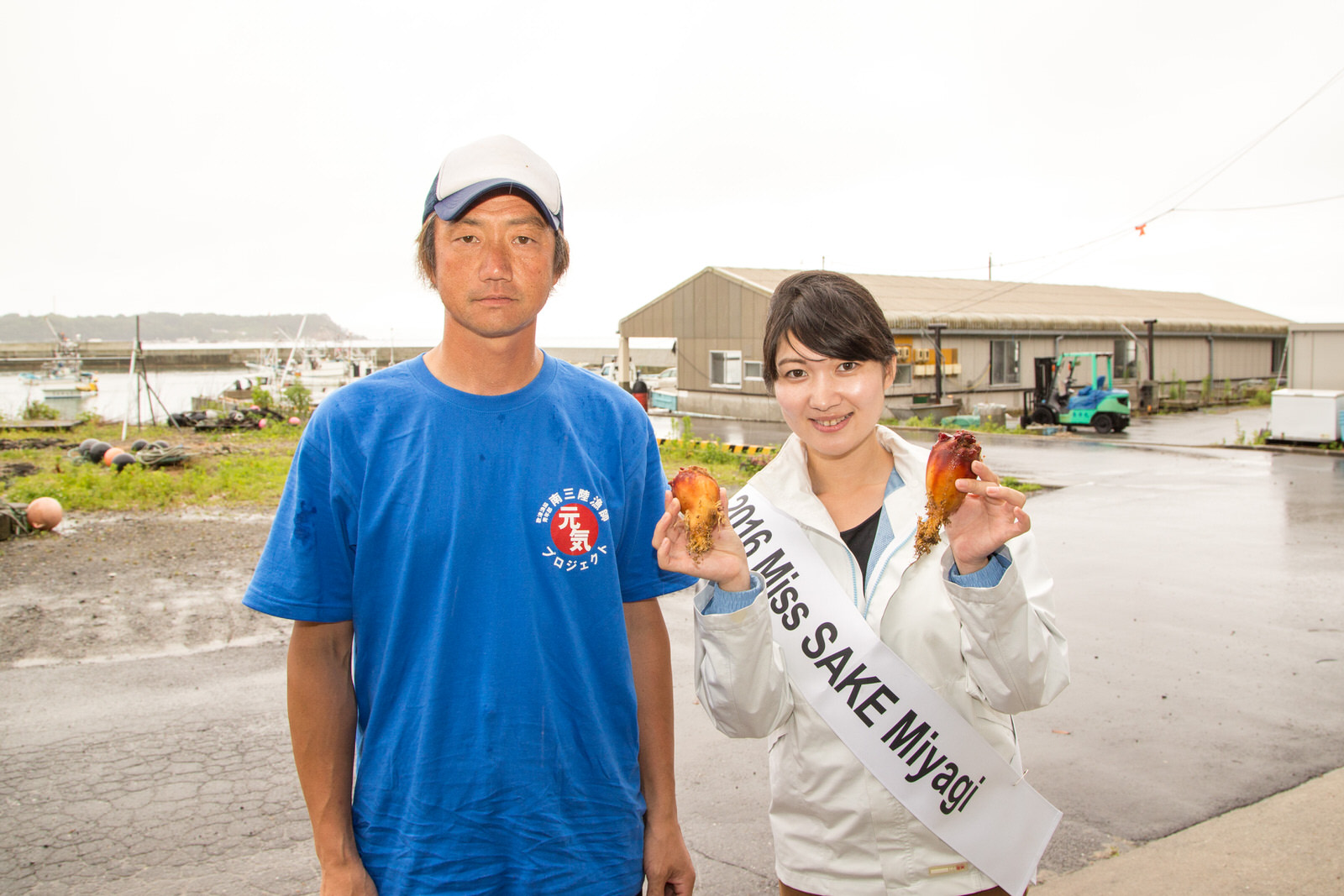 港近くで青いTシャツの男性と2016 Miss SAKE Miyagiのたすきをかけた女性が並び、ホヤを手に笑顔を見せている写真