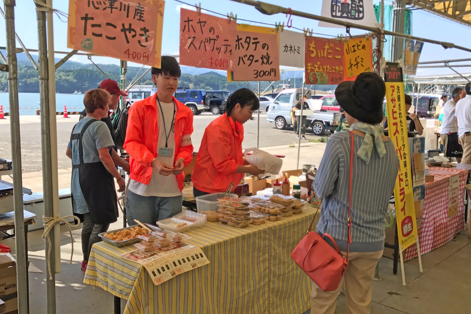 港の近くで行われているイベントでたこ焼きやホヤ、ホタテを使った料理の屋台をオレンジのジャンバーを着た学生二人が運営しており、女性客が屋台に立ち寄っている様子の写真いる
