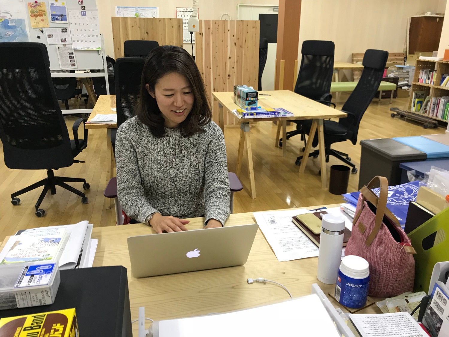 木製の机や本棚が並ぶオフィスの中で、グレーのセーターを着た女性がMacBookのノートパソコンで作業をしている写真