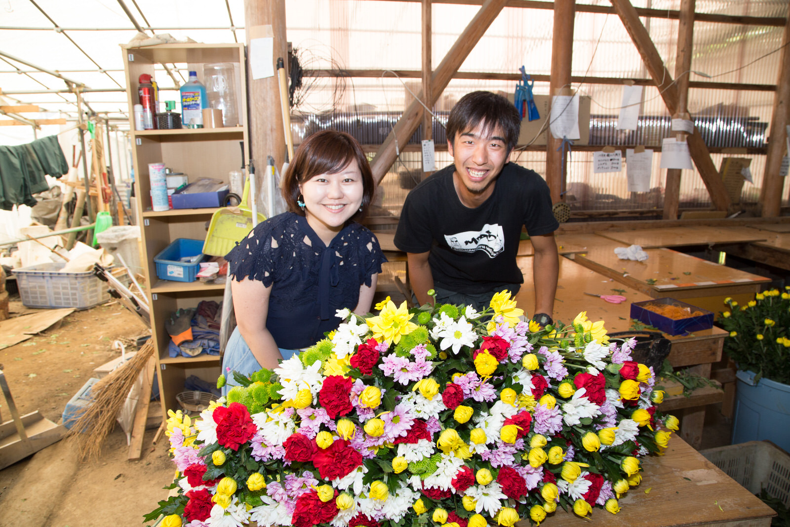 作業台や様々な道具が置かれた作業場で、色とりどりの花が山のように乗っている台の後ろに並んで立っている、リポーターの女性と浅野さんの写真