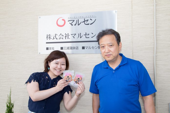 株式会社マルセンのシルバーの看板が取り付けられた壁の前で、三浦さんと、個包装のタコ天を持ったリポーターの女性が並んで立っている写真