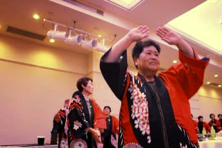 赤と黒の法被を着た女性たちが会場内で輪になり、楽しそうに手を上げて盆踊りをしている様子の写真