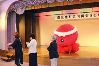 ステージ上に赤いタコのオクトパス君が立っており、その下で3人の女性参加者が手拍子をして踊っている写真