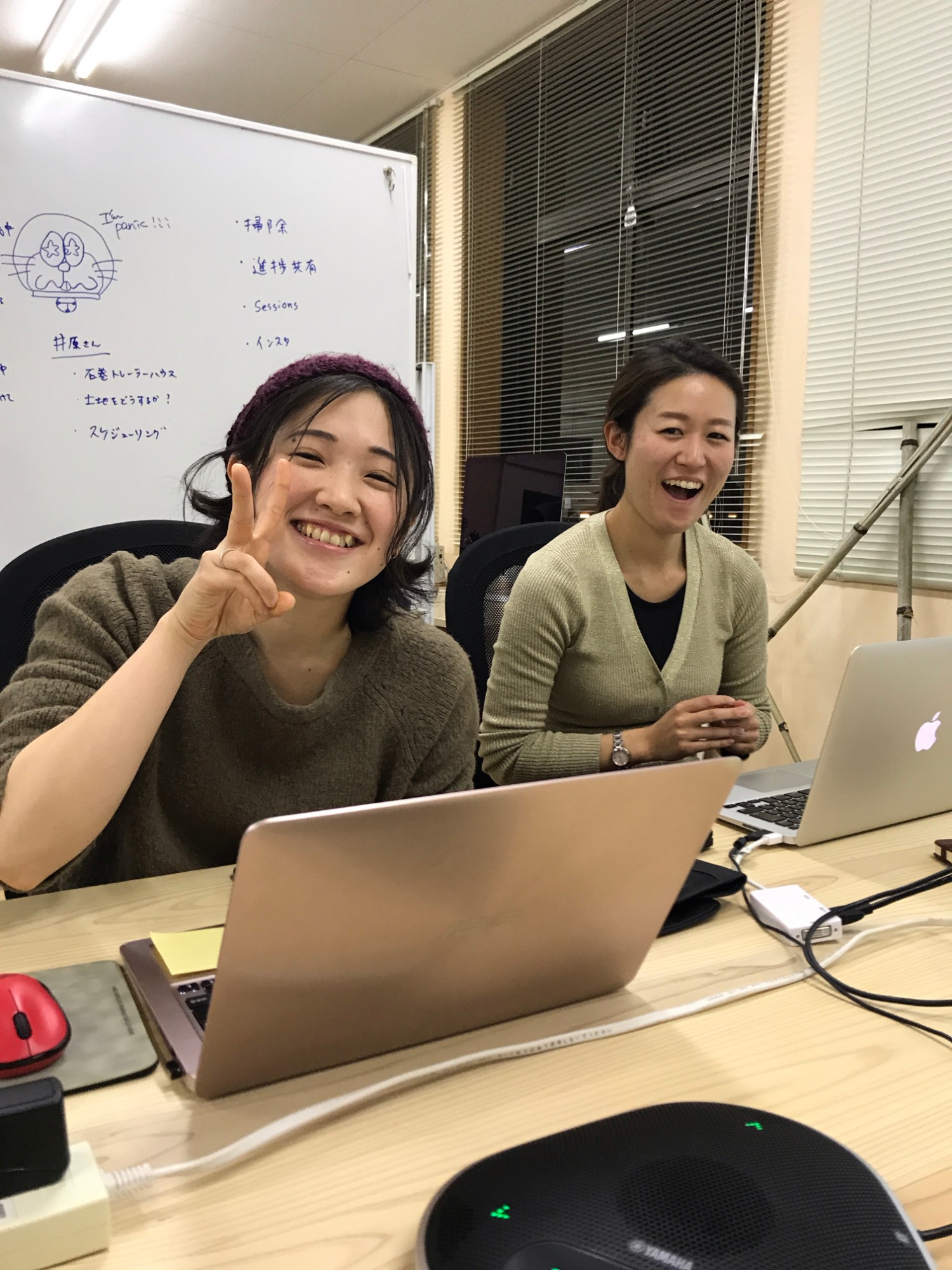 白いボードが背景にあり、机の上に並んだノートパソコンに向かい、1人はピースサインをしながら笑顔で、もう1人は口を開けて笑っている2人の女性の写真