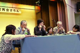 ステージの上で水色のクロスがかけられた机を囲んだ2人の男性と女性が、ハーモニカ演奏を披露している様子の写真