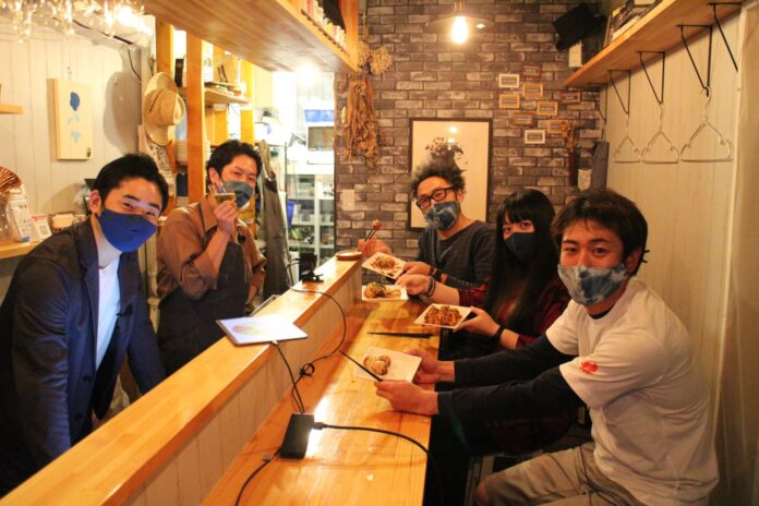 YouTubeでライブ配信された南三陸移住トークイベントの収録時に、地域おこし協力隊のメンバー3人がタコ焼きを手に笑顔で談笑しており、店主ともう一人の関係者がカウンター越しに交流している様子の写真